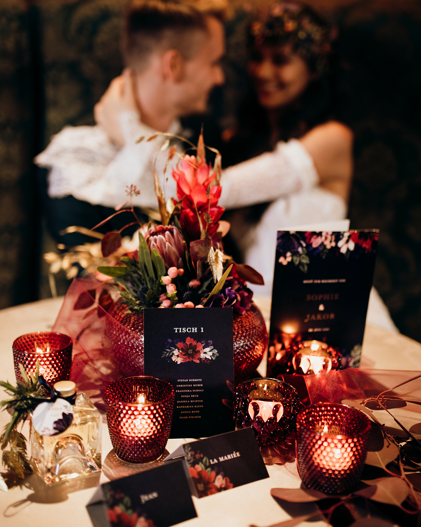 Im Vordergrund ist eine Tischkarten im Boho-Look auf der Hochzeitstafel platziert. Im Hintergrund schaut sich das Brautpaar verliebt an.
