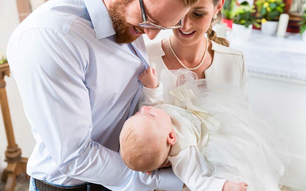 Baby im Taufkleid hält seinen Papa am Hemd fest, während die Mama lachend daneben steht.
