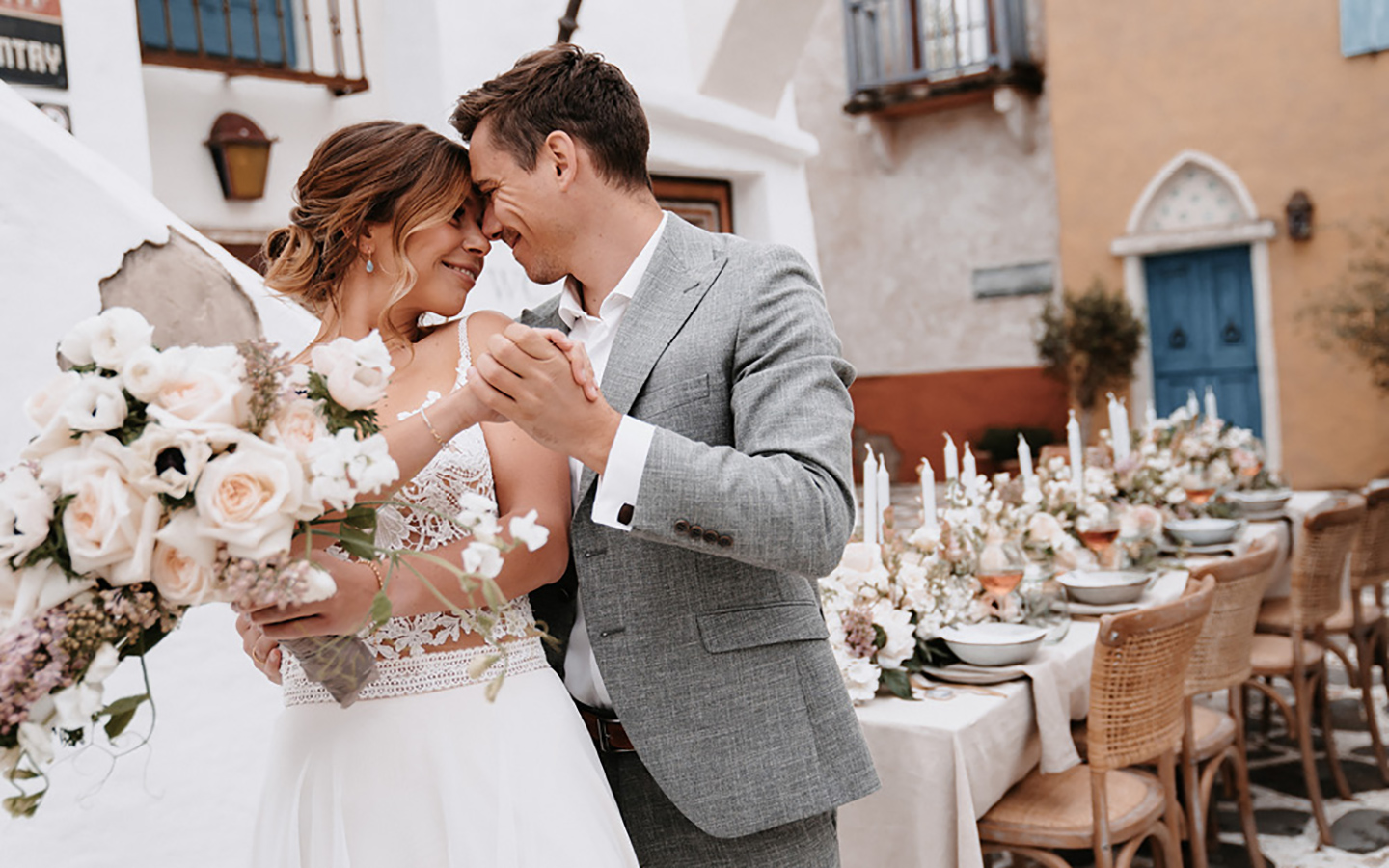 Braut und Bräutigam sehen sich tief in die Augen und strahlen über das ganze Gesicht. Der Blumenstrauß der Braut in hellen Tönen wird von ihr in der Hand gehalten. Im Hintergrund ist der Hochzeitstisch zur mediterranen Hochzeit zu sehen.