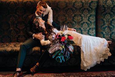 Die Braut liegt auf den Knien des Bräutigams. Die Beiden schauen sich verliebt an. Die Frau trägt ein boho angehauchtes Brautkleid aus Spitze und der Mann einen moosgrünen Anzug. In der Hand hält die Braut einen üppigen Brautstrauß in bunten Farben.