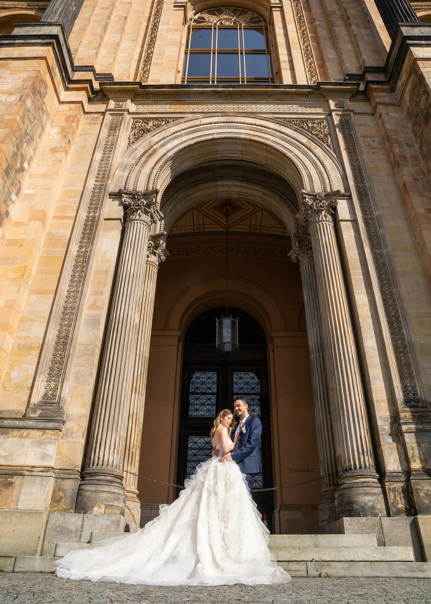 Das Hochzeitspaar bestehend aus Braut und Bräutigam steht vor einer beeindruckenden Architektur, in der die Trauung stattfindet. Vor der imposanten Tür posieren sie für Fotos.