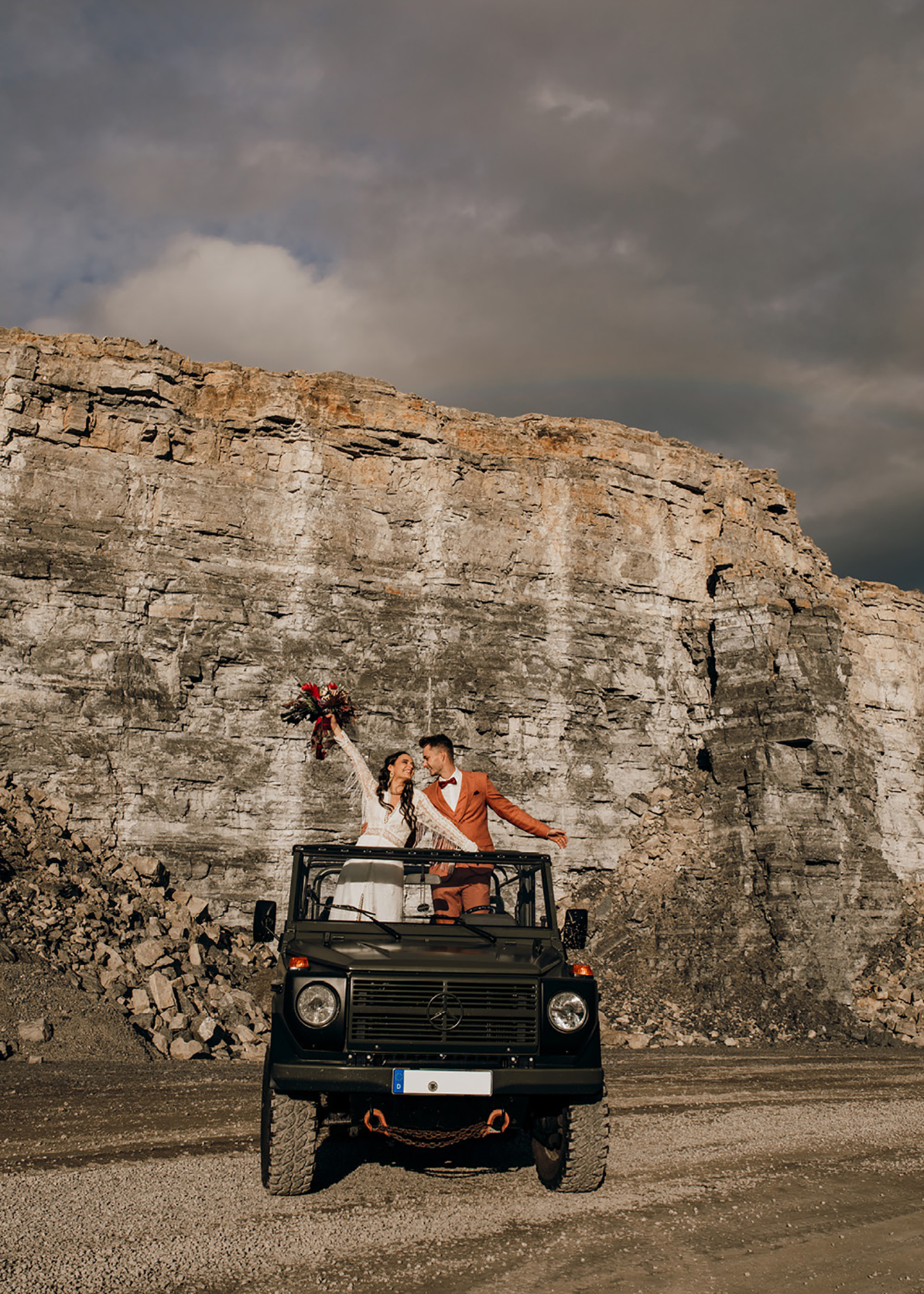 Die beeindruckende Kulisse des Shootings - ein Steinbruch, im Vordergrund das Brautpaar auf dem Geländewagen, dunkle Wolken und ein Regenbogen hinter ihnen
