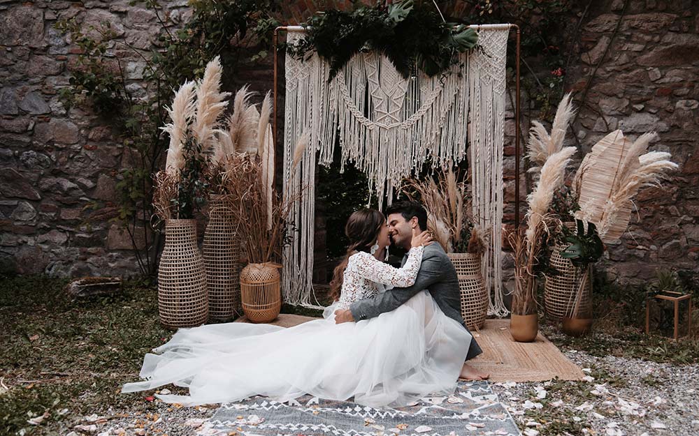 Boho-Greenery Hochzeit im Freien: Das verliebte Brautpaar wird vor der romantischen Boho Dekoration aus Pampasgras und Makramee fotografiert.
