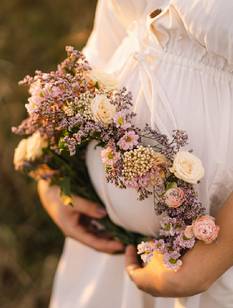 Bildausschnitt: Hochschwangere Frau in weißem Kleid hält Blumenkranz vor ihrem Bauch