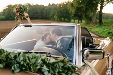 Frisch vermähltes Hochzeitspaar sitzt sich küssend im einem alten Oldtimer. Das Hochzeitsauto ist festlich geschmückt und die Braut streckt ihren Brautstrauß in die Luft.