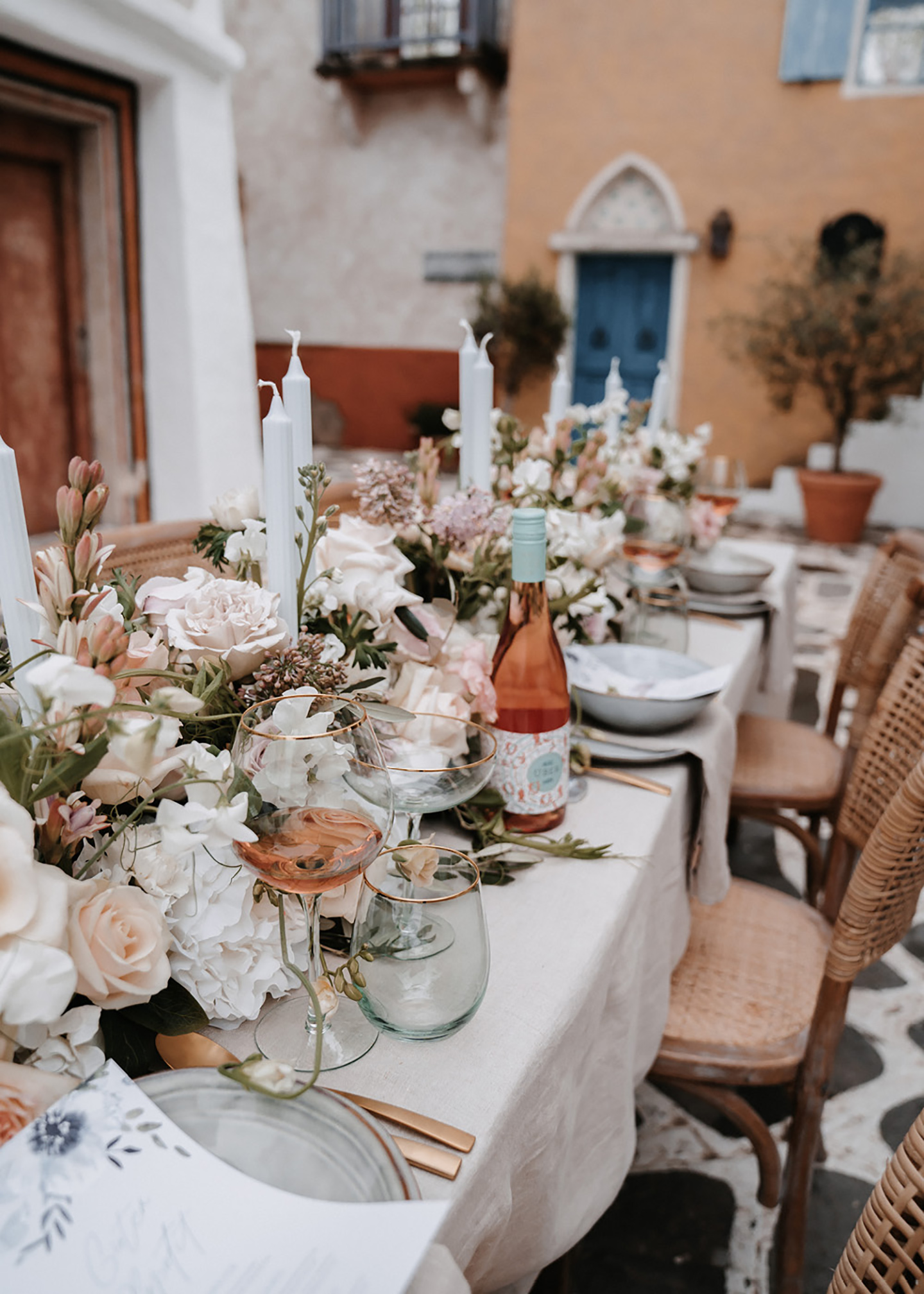 Der angerichtete mediterrane Hochzeitstisch erstrahlt mit einer Pracht aus Blumen und Dekoration. Das Besteck ist rosegold und passt zu den beige-pastellorangenen Häusern im Hintergrund.