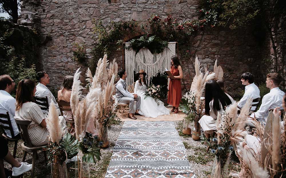 Während der Trauung blickt die Braut ihrem Ehemann glücklich in die Augen. Die Gäste schauen gespannt bei der Trauung zu. 