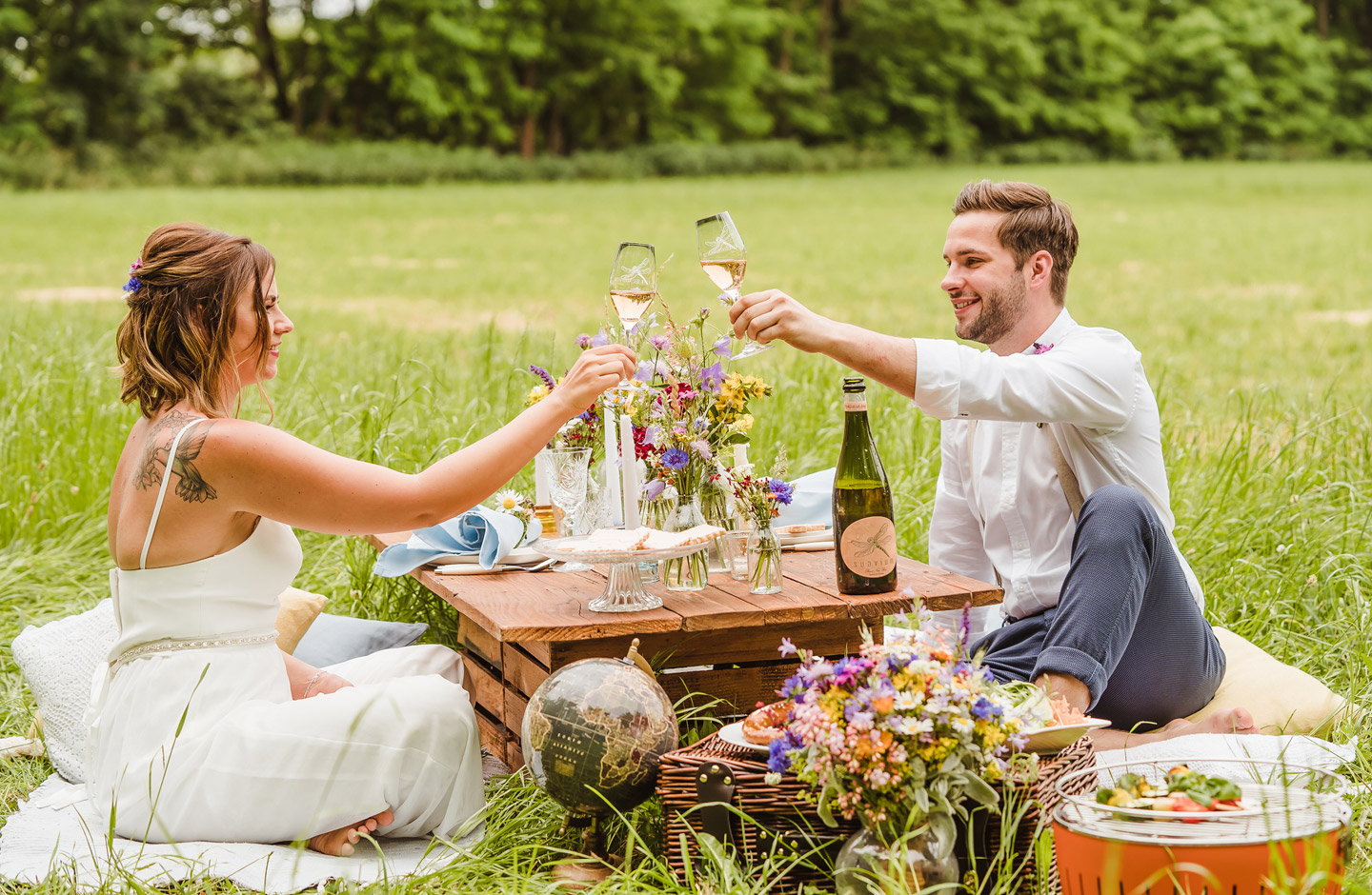 Hochzeitspaart stößt mit Sekt am Picknicktisch auf einer Wiese auf ihre Hochzeit an.