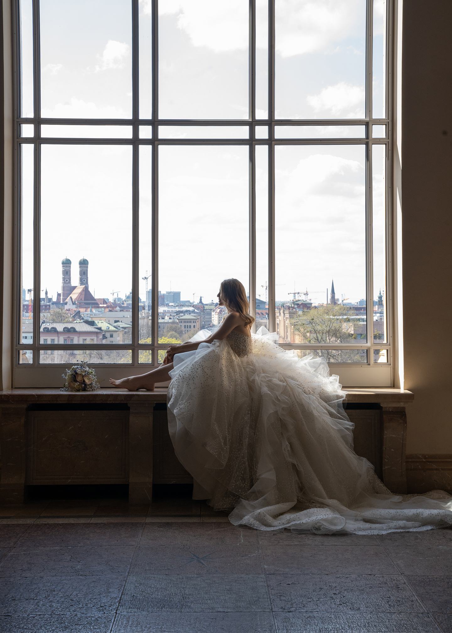 Die Braut sitzt in ihrem pompösen Prinzessinnenkleid passend zum Stil der traditionellen Hochzeitvor einem großen Fenster in der Location und posiert für Fotos.