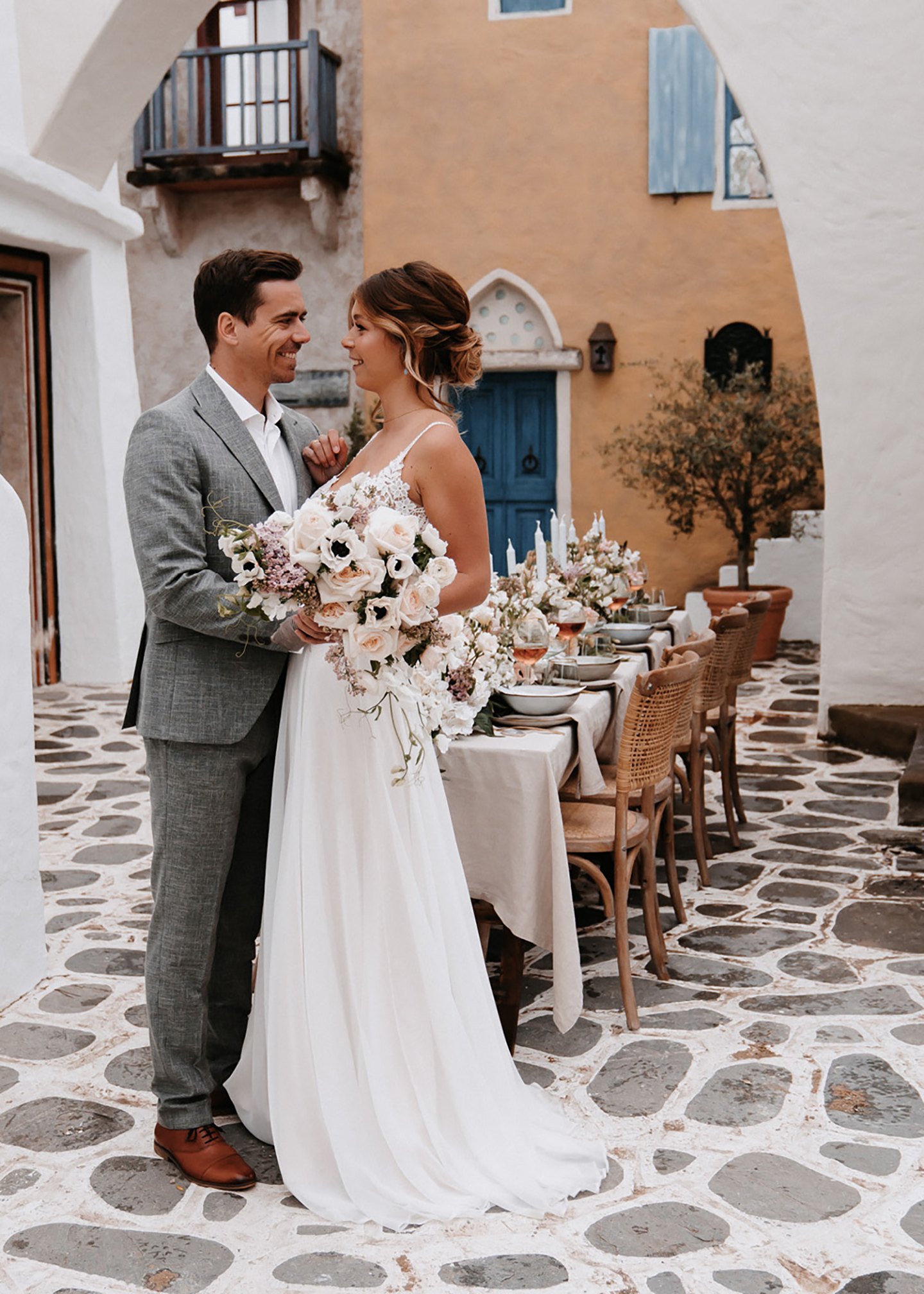 Das glückliche Hochzeitspaar aus Braut und Bräutigam steht vor der Location mit gedecktem Hochzeitstisch und schaut sich verliebt an.