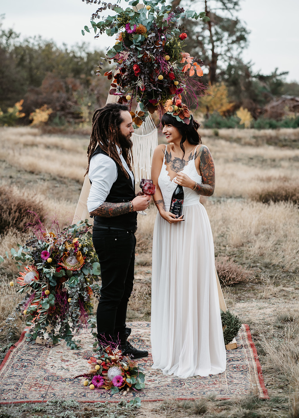 Das Brautpaar steht vor dem mit bunten Blumen geschmückten Traubogen auf der Wiese aus hellen Gräsern in der Hochzeitslocation.