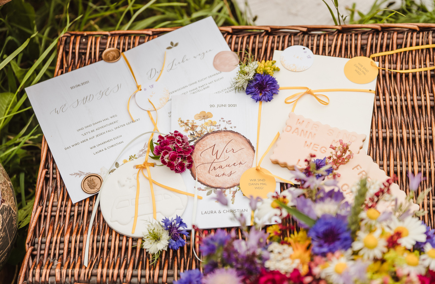Hochzeitseinladung zur Camper Hochzeit liegt auf einer geflochtenen Holz-Kiste und ist dekoriert mit Wildblumen.