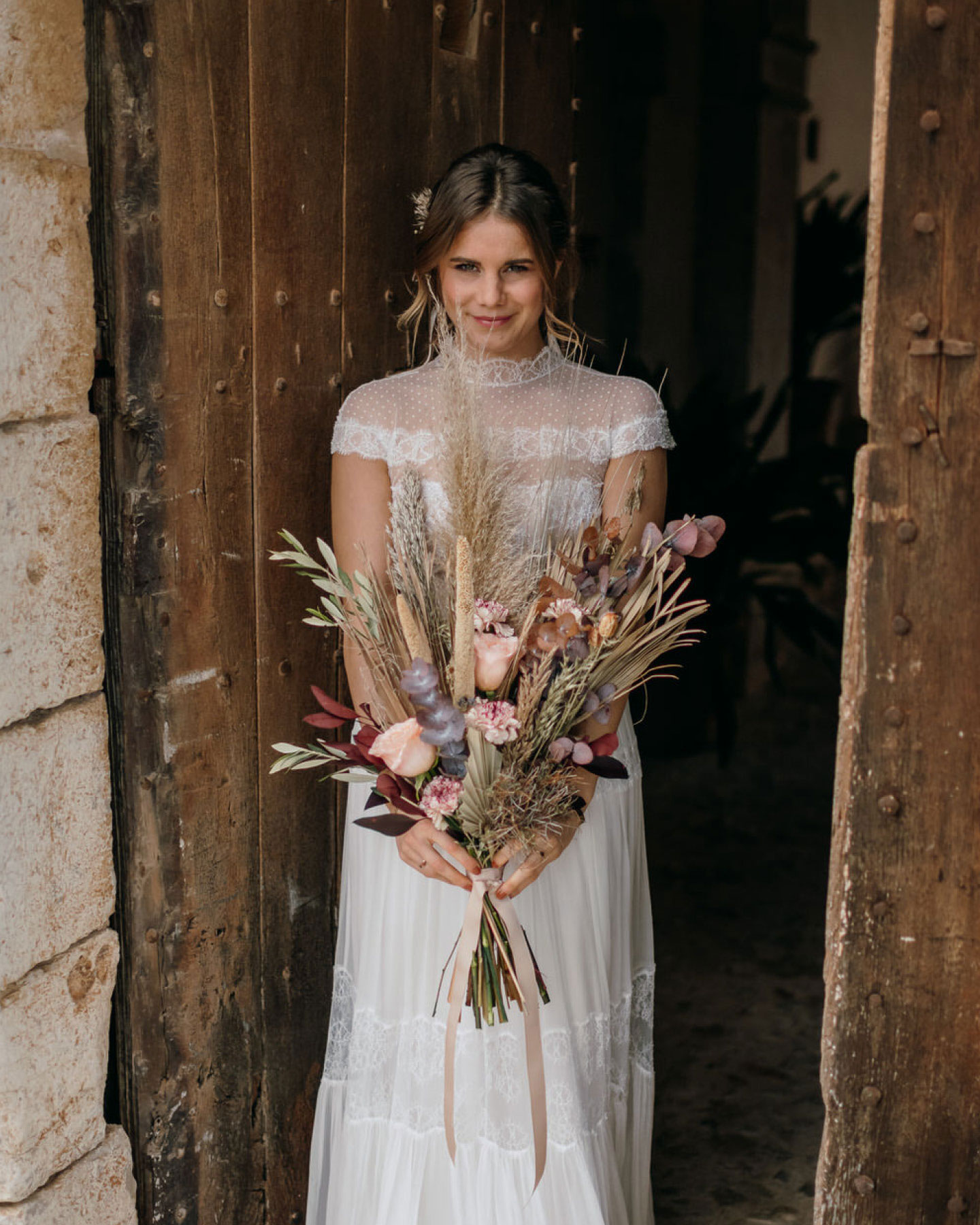 Braut tritt aus alter Scheune und hält einen Brautstrauß aus Trockenblumen.