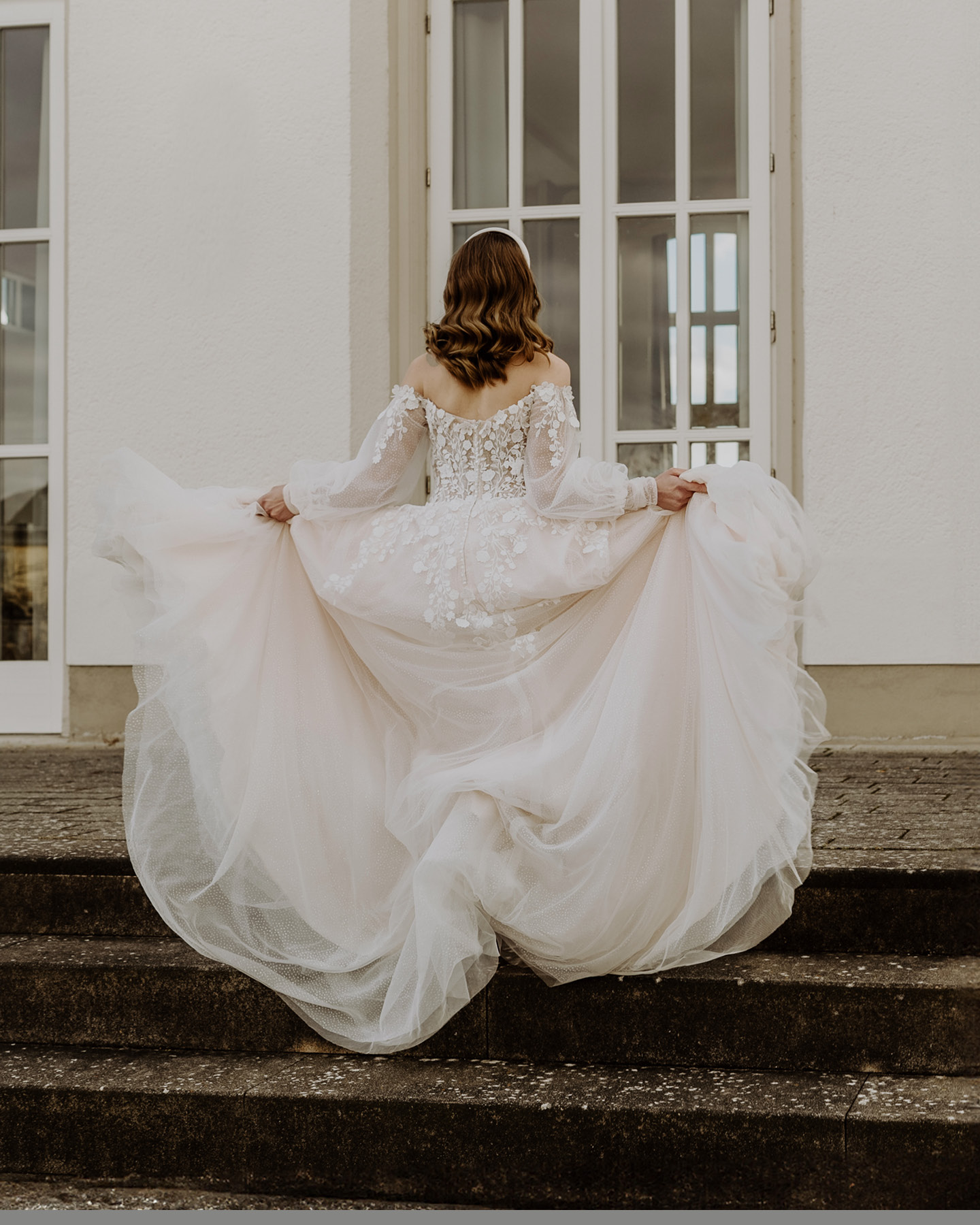 Braut hält Brautkleid mmit weitem Rock hoch und geht die Stufen zur Location hoch